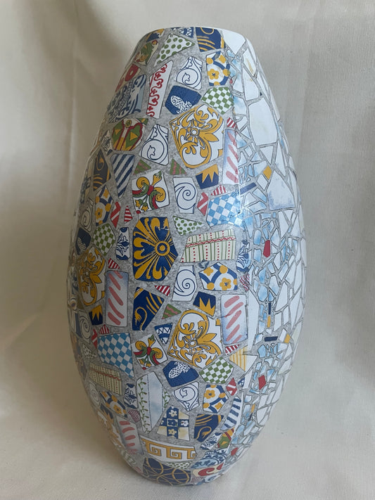 A tall, rounded ceramic vase covered in a colorful mosaic pattern of blue, yellow, red, white, and gray decorative tiles, set against a neutral beige fabric backdrop.