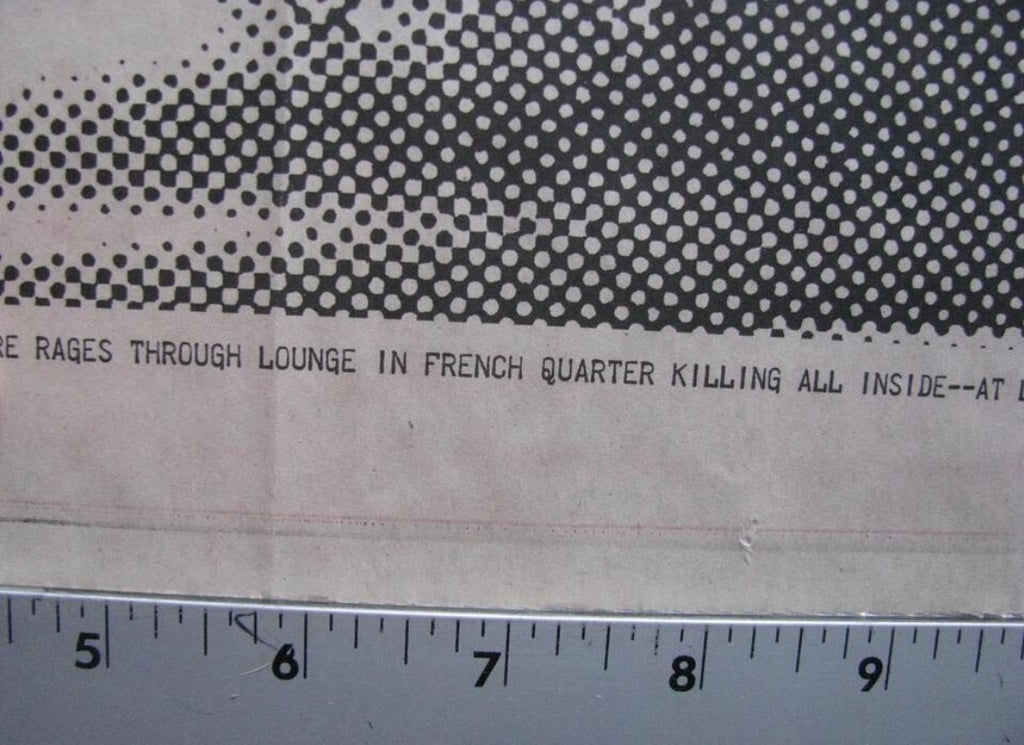 A close-up of a printed paper item with a black halftone dot pattern across the top and partial black text reading 'RAGES THROUGH LOUNGE IN FRENCH QUARTER KILLING ALL INSIDE--AT D', with a metal ruler along the bottom edge for scale.