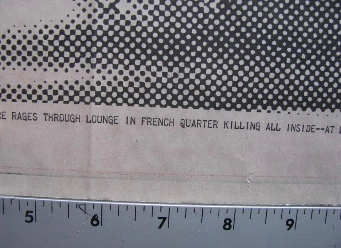 A close-up of a printed paper item with a black halftone dot pattern across the top and partial black text reading 'RAGES THROUGH LOUNGE IN FRENCH QUARTER KILLING ALL INSIDE--AT D', with a metal ruler along the bottom edge for scale.