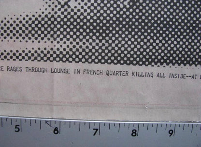 A close-up of a printed paper item with a black halftone dot pattern across the top and partial black text reading 'RAGES THROUGH LOUNGE IN FRENCH QUARTER KILLING ALL INSIDE--AT D', with a metal ruler along the bottom edge for scale.