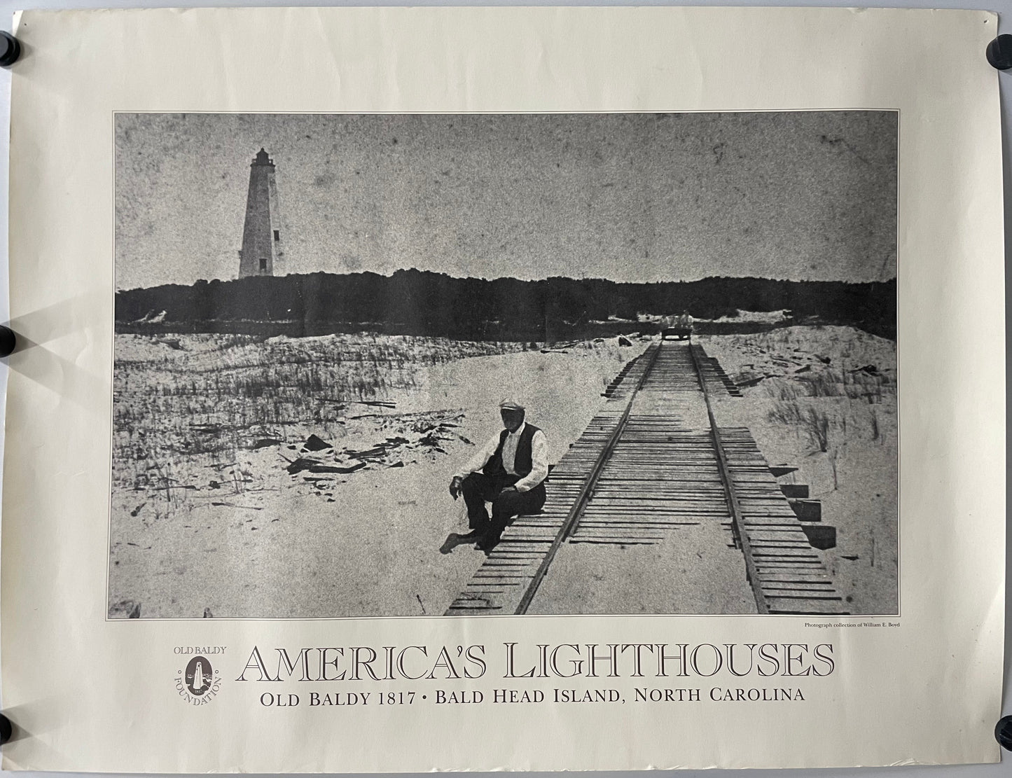 original and vintage, America’s Lighthouses  - Old Baldy Foundation, exhibition poster black and white image of a man sitting on a dock with a  lighthouse in the background on Bald Head Island.