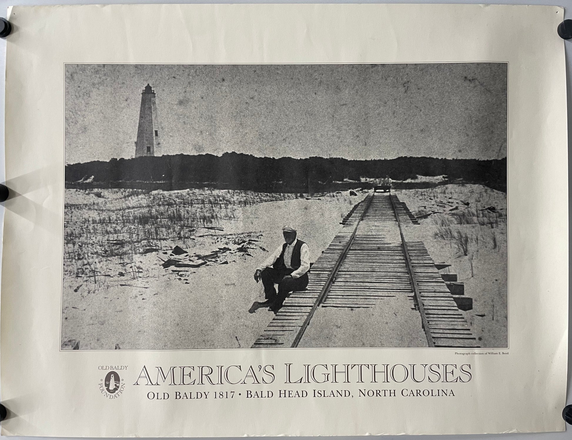 original and vintage, America’s Lighthouses  - Old Baldy Foundation, exhibition poster black and white image of a man sitting on a dock with a  lighthouse in the background on Bald Head Island.