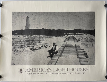 original and vintage, America’s Lighthouses  - Old Baldy Foundation, exhibition poster black and white image of a man sitting on a dock with a  lighthouse in the background on Bald Head Island.
