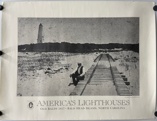original and vintage, America’s Lighthouses  - Old Baldy Foundation, exhibition poster black and white image of a man sitting on a dock with a  lighthouse in the background on Bald Head Island.