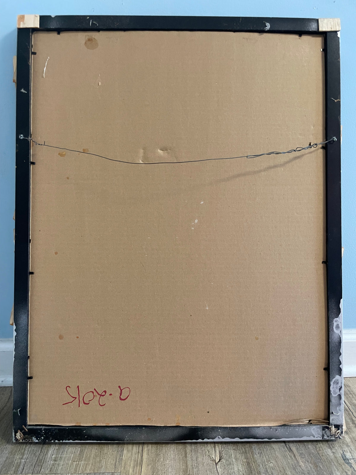 Back of a framed painting with visible wear and signature, against a blue wall.