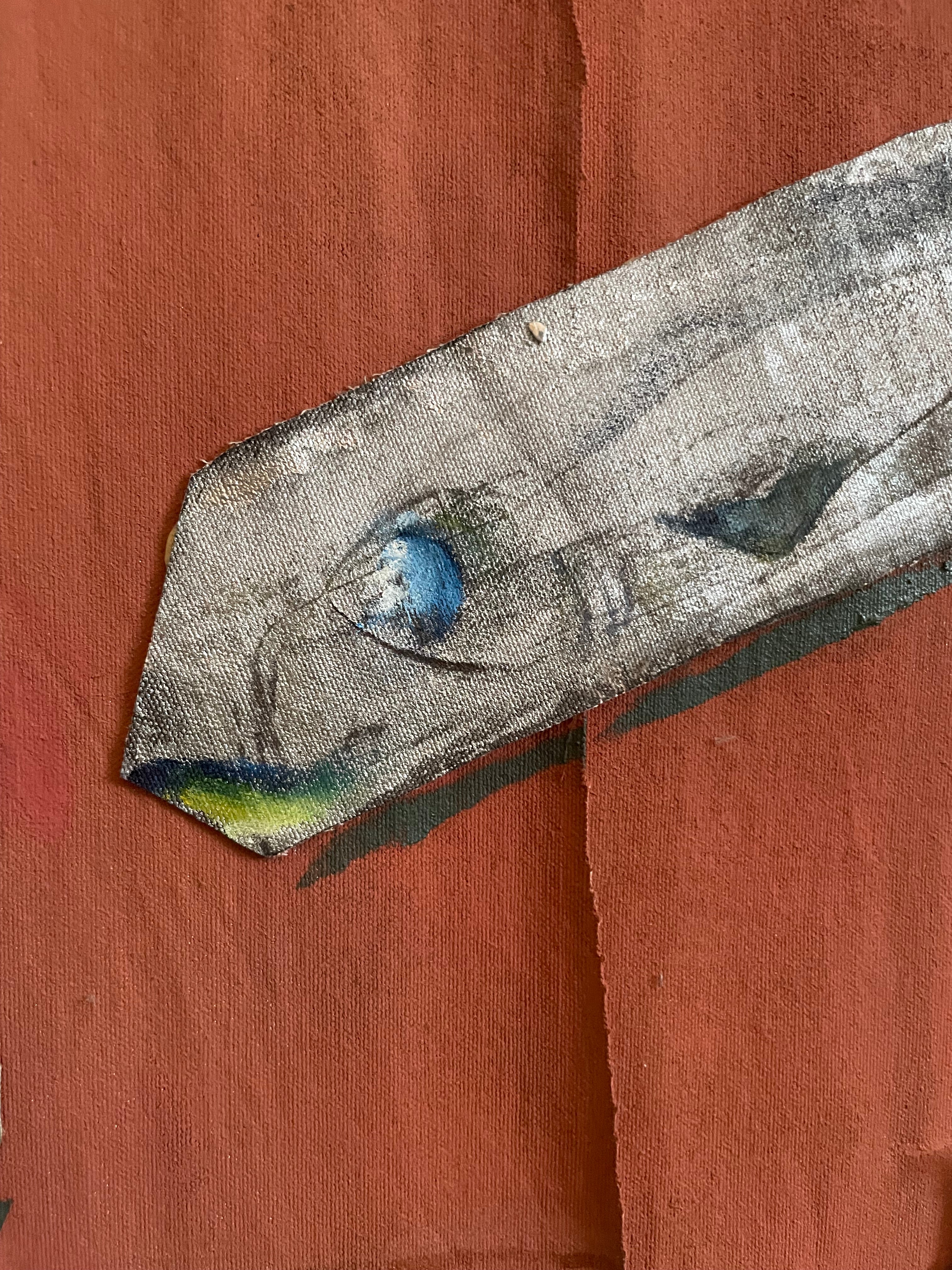 A close-up of a textured gray fabric or canvas patch with a painted eye motif and small blue, green, and yellow accents, laid diagonally on a reddish-brown background.