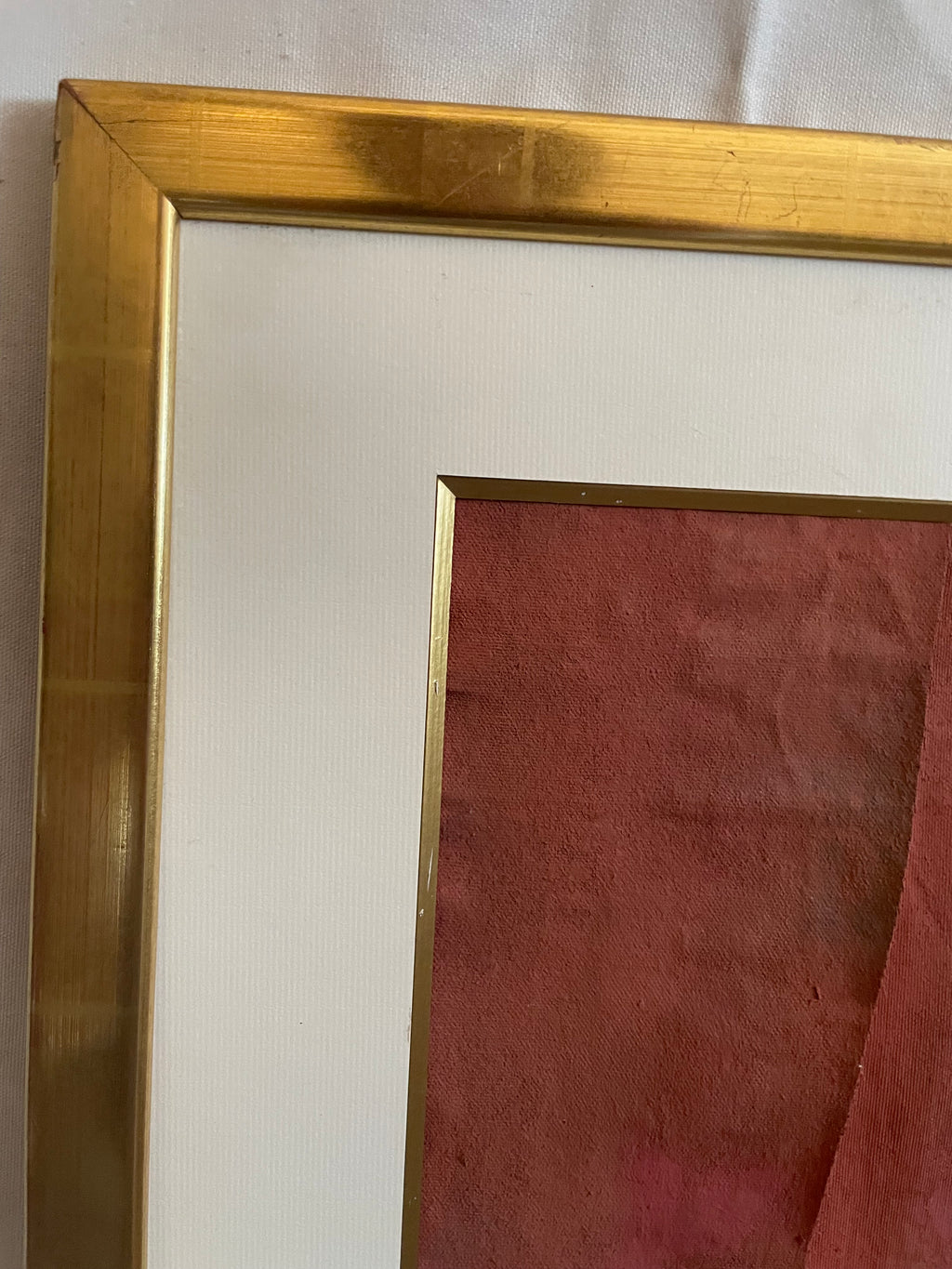 Close-up of a framed art print showing a gold metal frame, white mat board, and a reddish-brown textured paper background.
