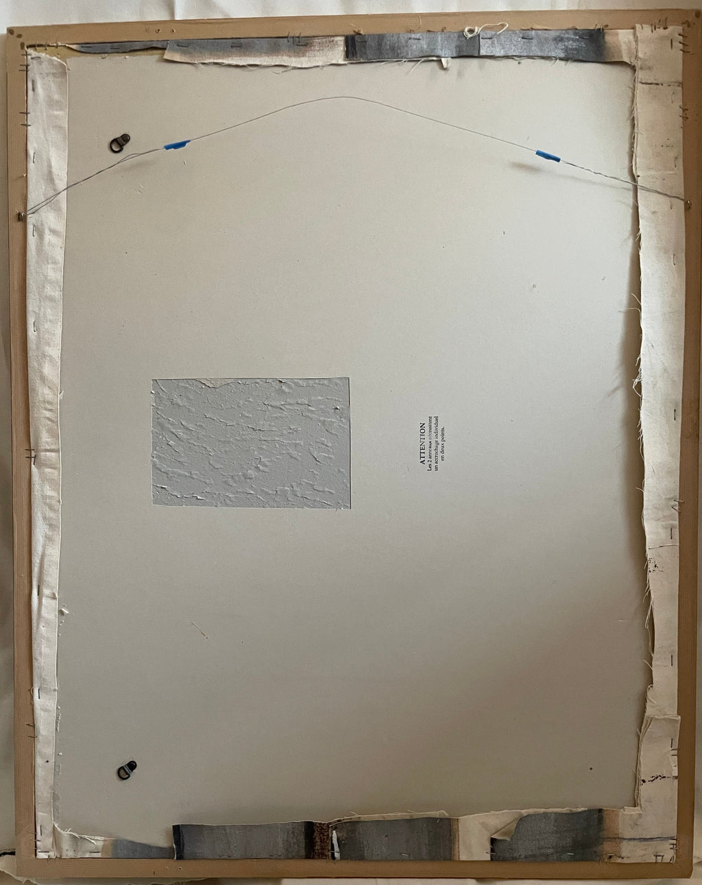 The back side of 'Apple of My Eye' (La Manzanita), showing a light wood frame, white backing board, mounting hardware, and a small central label on a white fabric background.