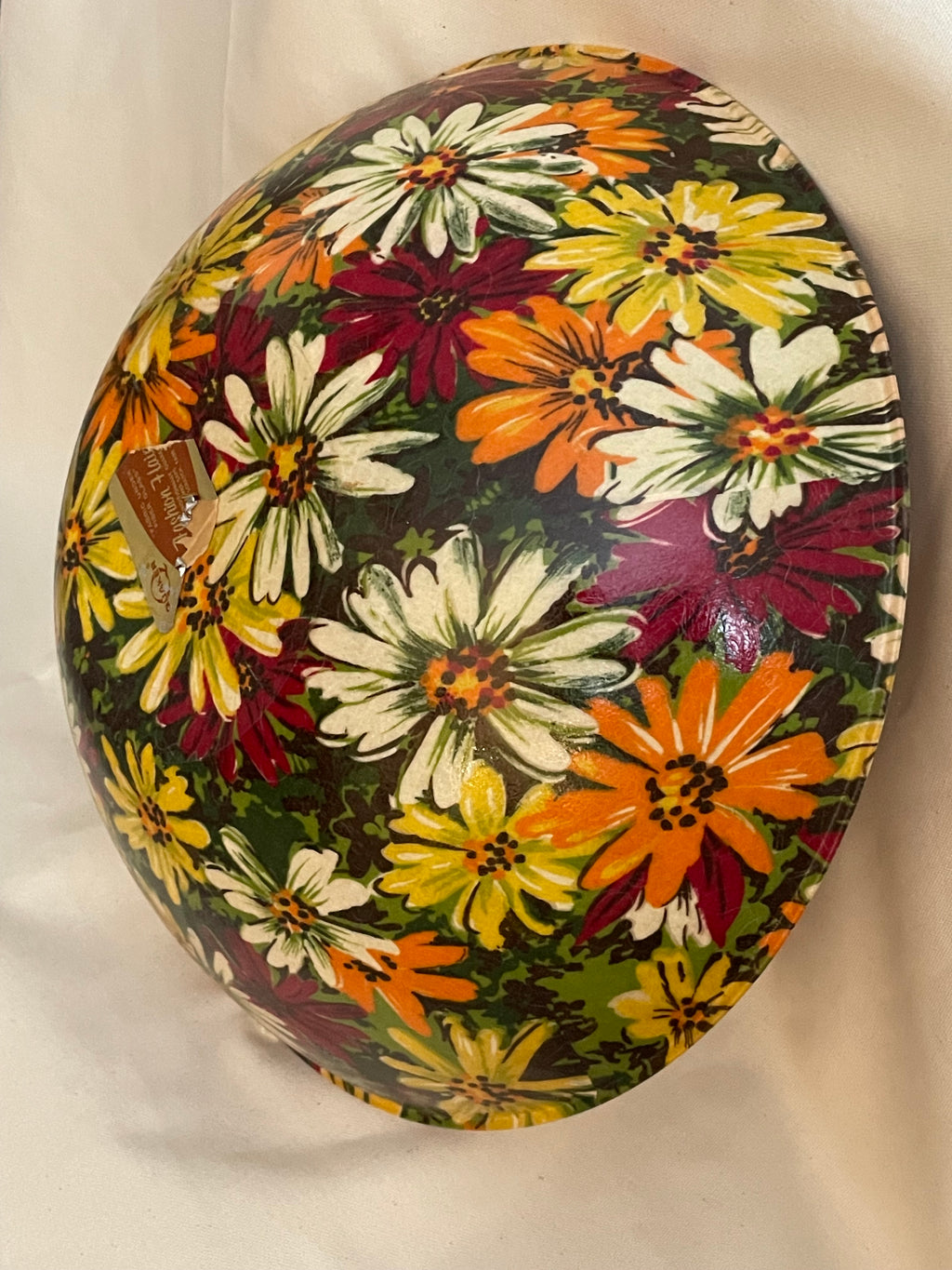 A round decorative bowl with a vibrant multicolor floral print featuring daisies in yellow, orange, white, and burgundy on a dark green background, photographed against a light beige fabric backdrop.