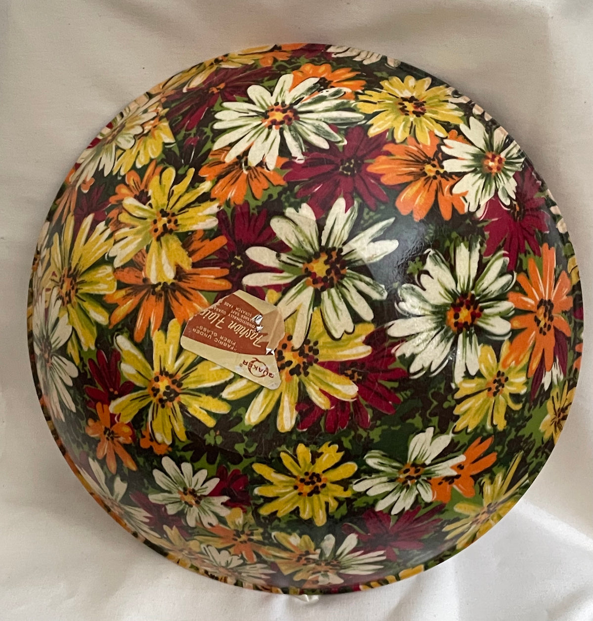 A round decorative bowl with a colorful floral print, featuring white, yellow, orange, and burgundy flowers on a dark background. A small paper tag is attached to the center.