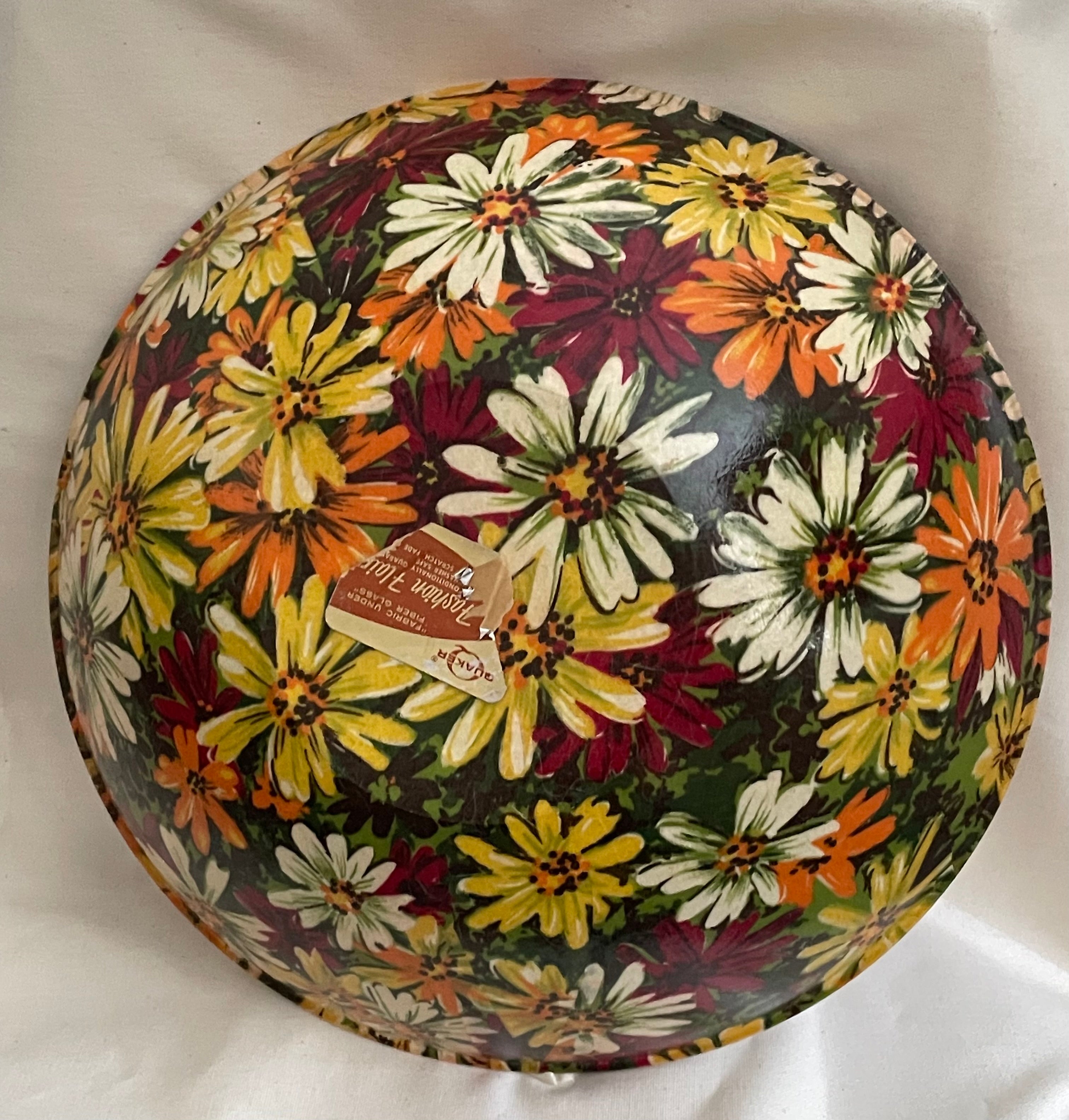 A round decorative bowl with a colorful floral print, featuring white, yellow, orange, and burgundy flowers on a dark background. A small paper tag is attached to the center.