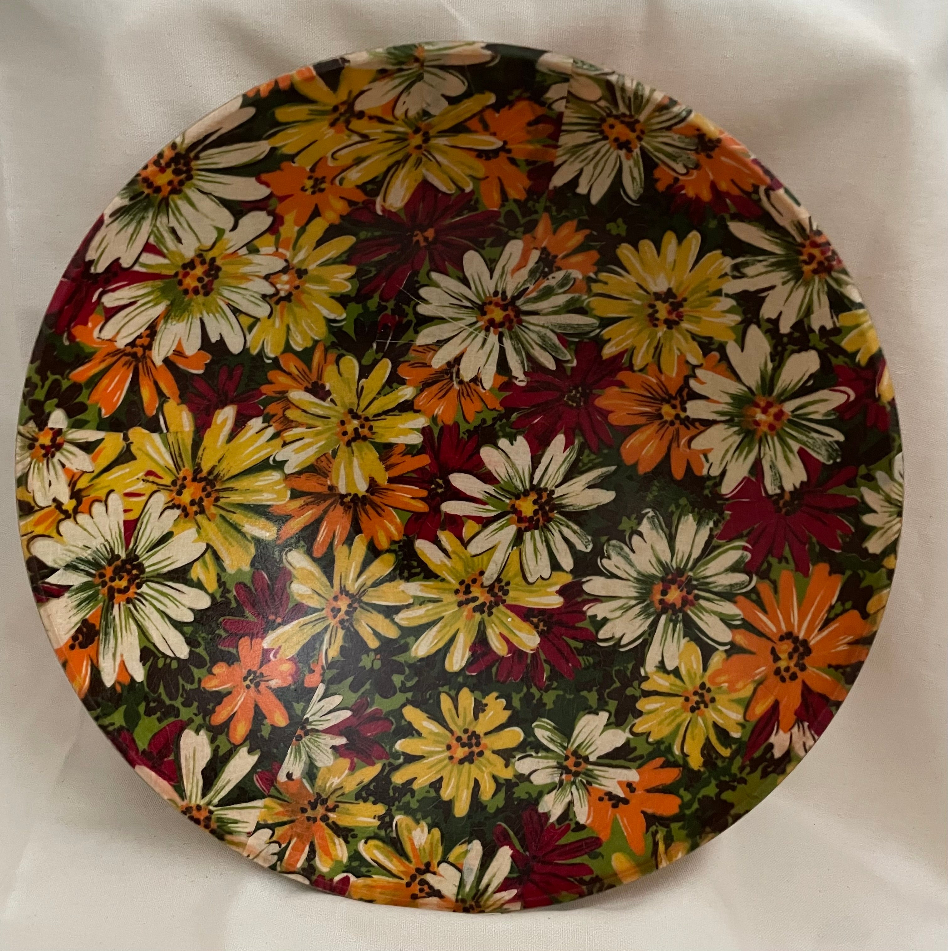 A round decorative plate featuring a dense floral pattern of daisy-like flowers in yellow, orange, white, and burgundy on a dark green background, photographed against a white fabric backdrop.