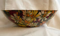 A decorative fabric and fiberglass bowl with a colorful floral pattern featuring white, yellow, and orange flowers with green leaves on a dark background, set against a draped white fabric backdrop.