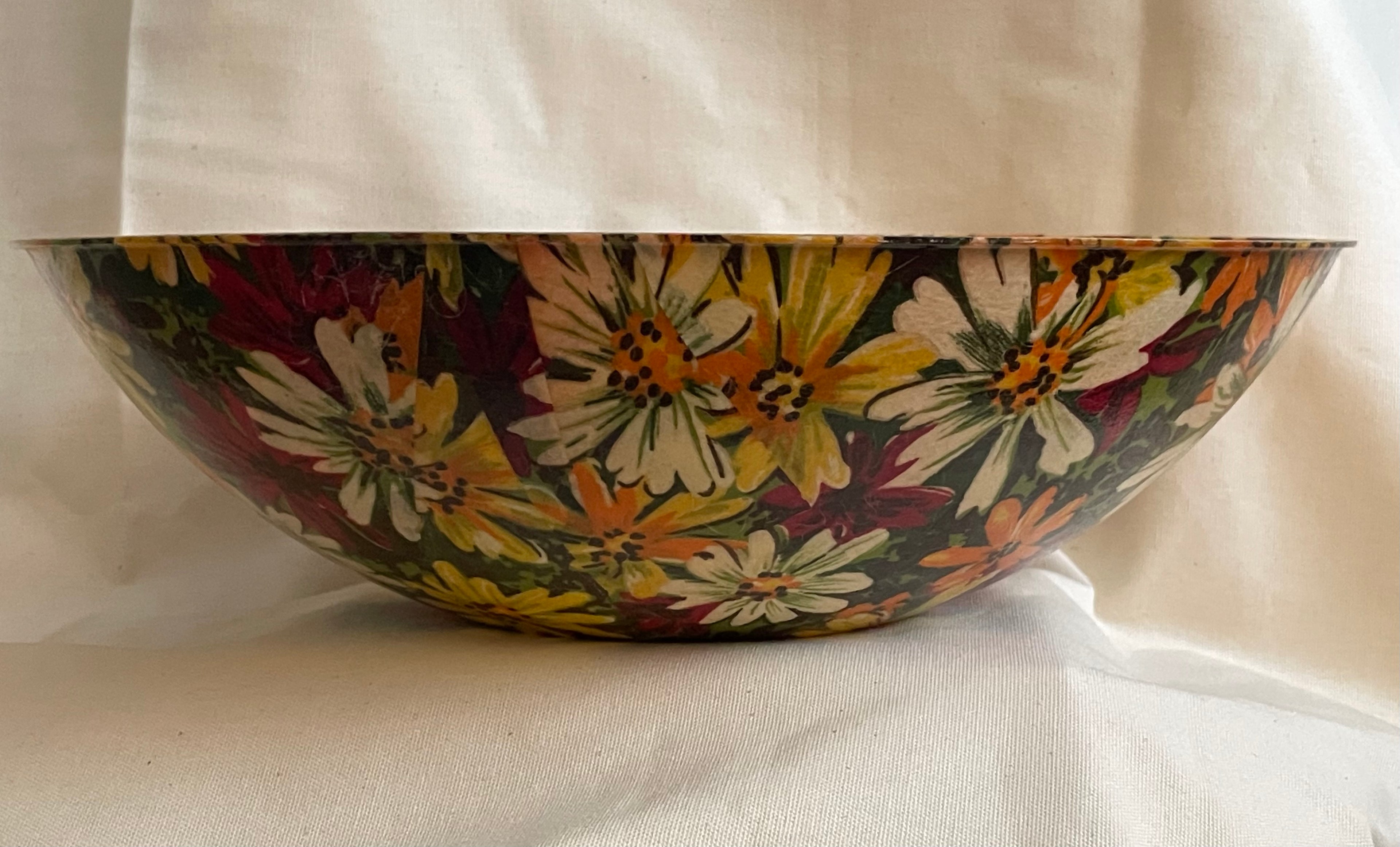 A decorative fabric and fiberglass bowl with a colorful floral pattern featuring white, yellow, and orange flowers with green leaves on a dark background, set against a draped white fabric backdrop.