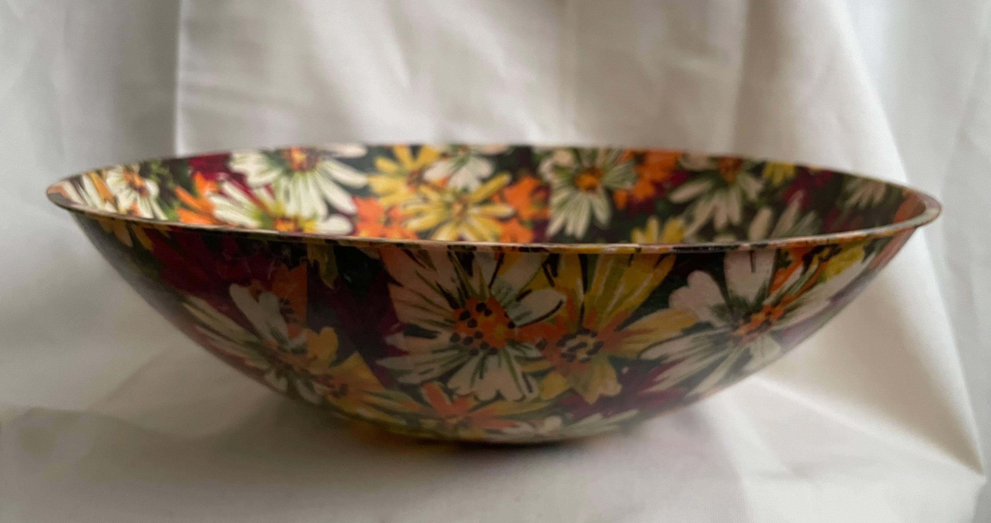 A wide, shallow decorative bowl with a dense floral pattern in orange, yellow, white, and burgundy on a dark background, set against a draped white fabric backdrop.
