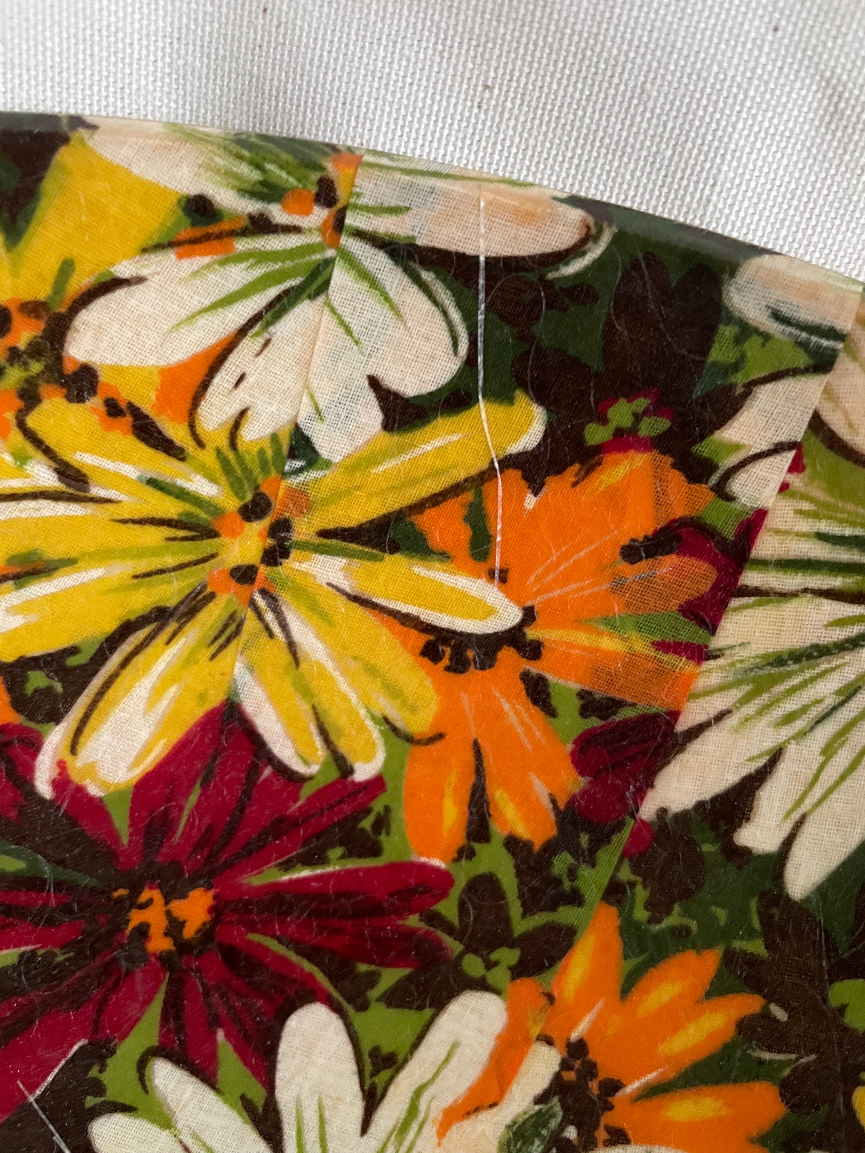 A close-up of a floral fabric with large daisy-like flowers in yellow, orange, burgundy, and white on a dark background, with a visible seam and slight fabric texture.