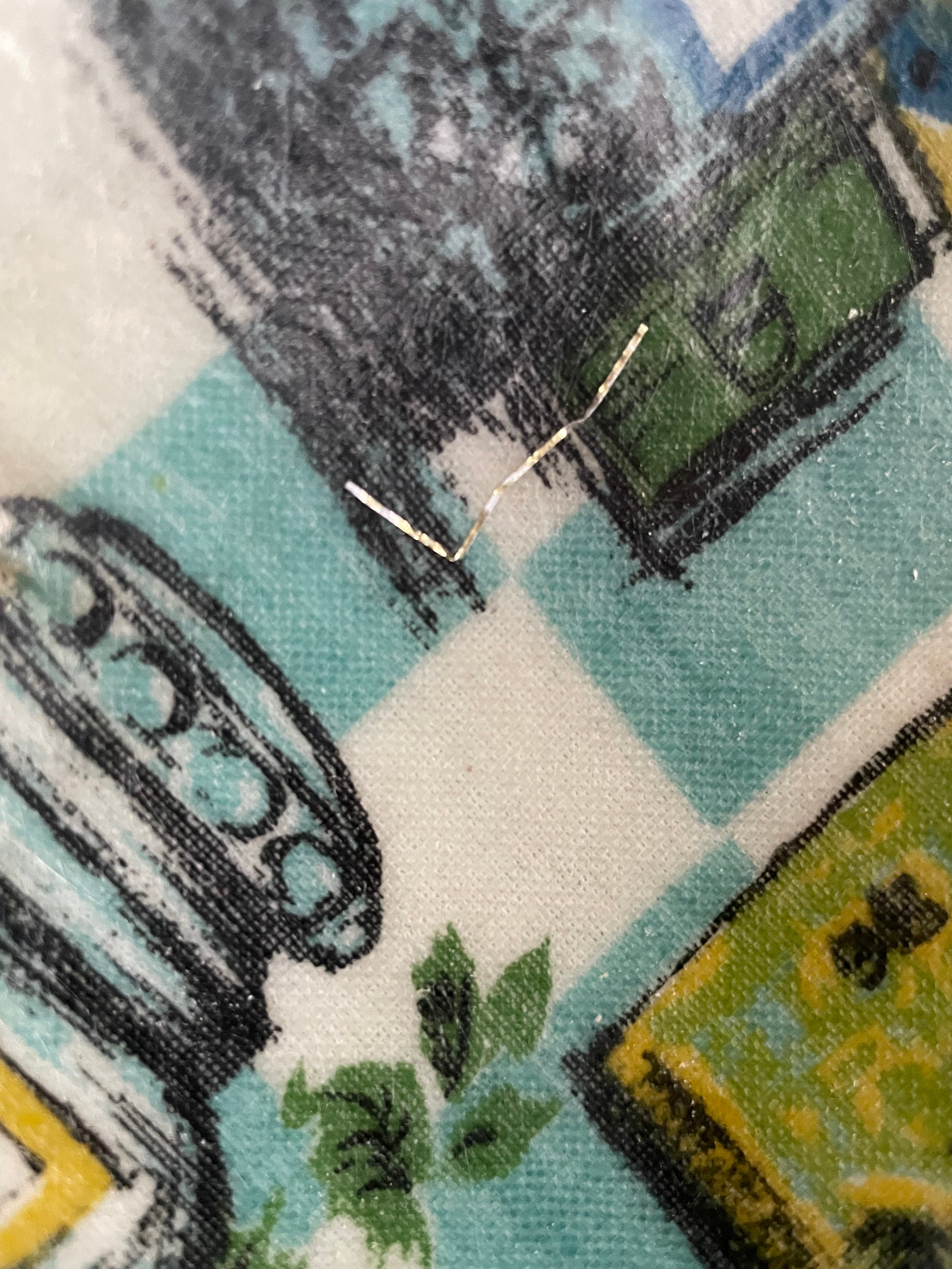 Close-up of a printed fabric with a vintage-style illustration in teal, white, black, green, and yellow, featuring a small metallic pin or needle-like mark near the center.