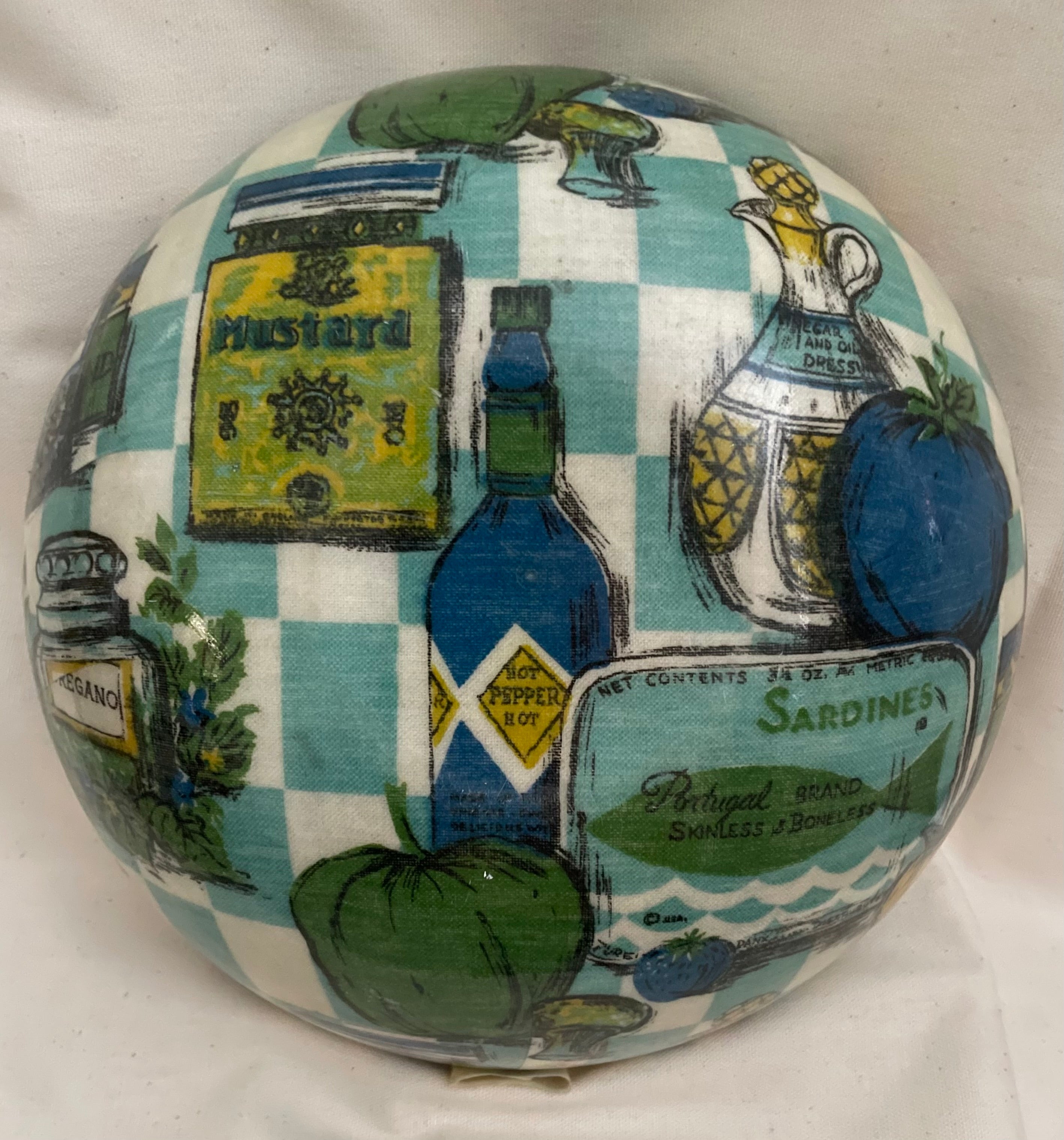 A decorative spherical object featuring a vintage-style printed collage of Mediterranean and kitchen motifs, including a green and white checkered pattern, illustrated mustard, pepper, olive oil, tomatoes, and a Sardines label.