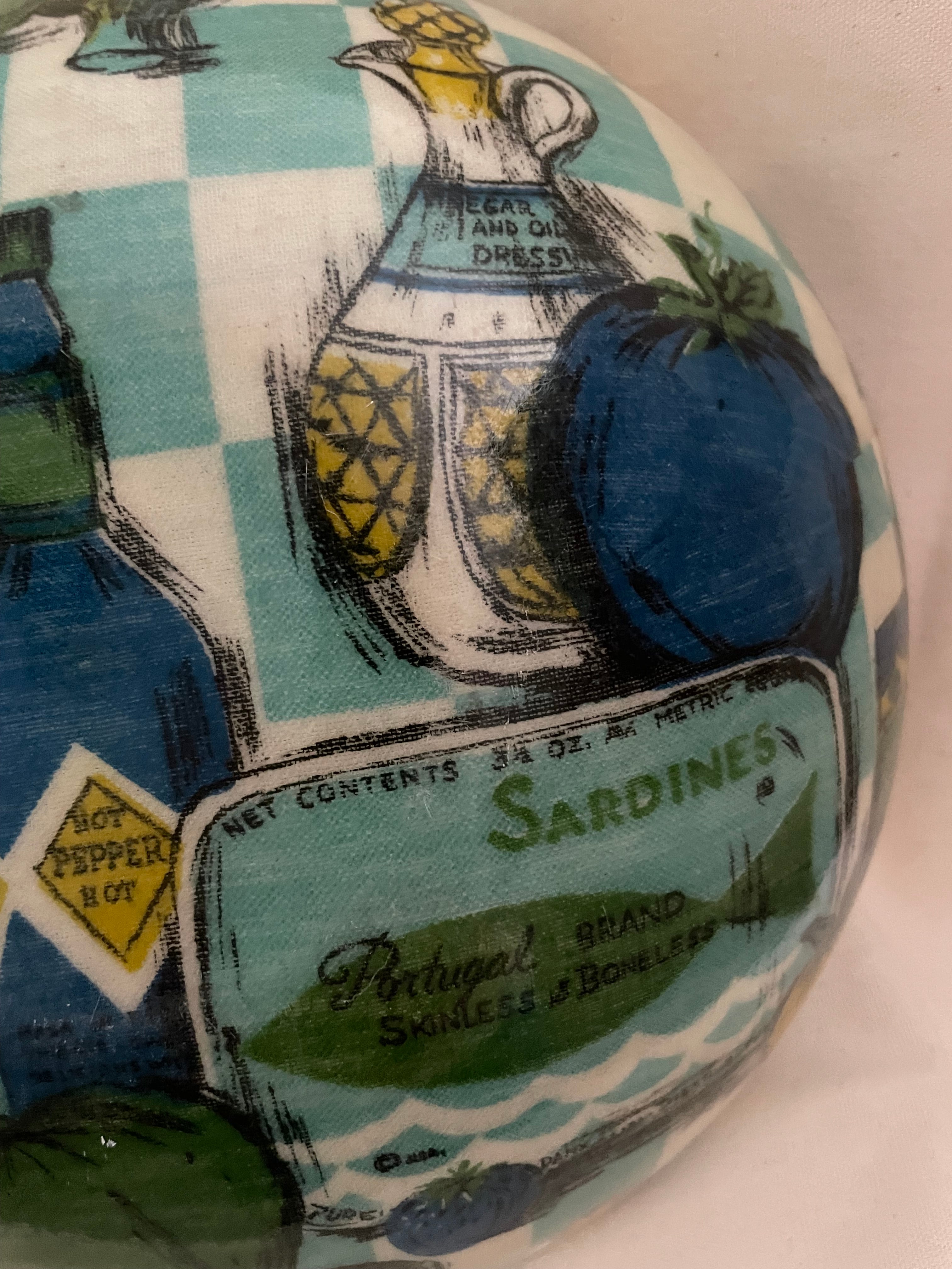 A close-up of a decorative ceramic container featuring a vintage-style Mediterranean sardine illustration with blue tomatoes, a white pitcher, and a green-and-white checkered pattern.