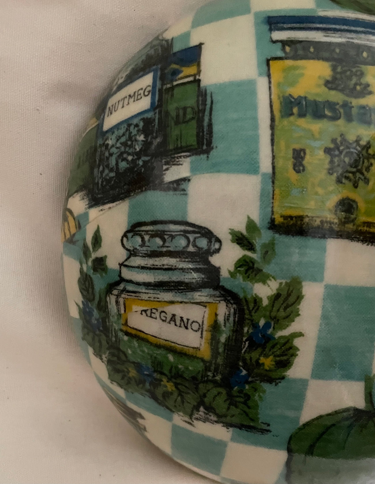 A close-up of a ceramic decorative object featuring a whimsical print of kitchen ingredients like nutmeg, mustard, and regano on a light blue and white checkered background.