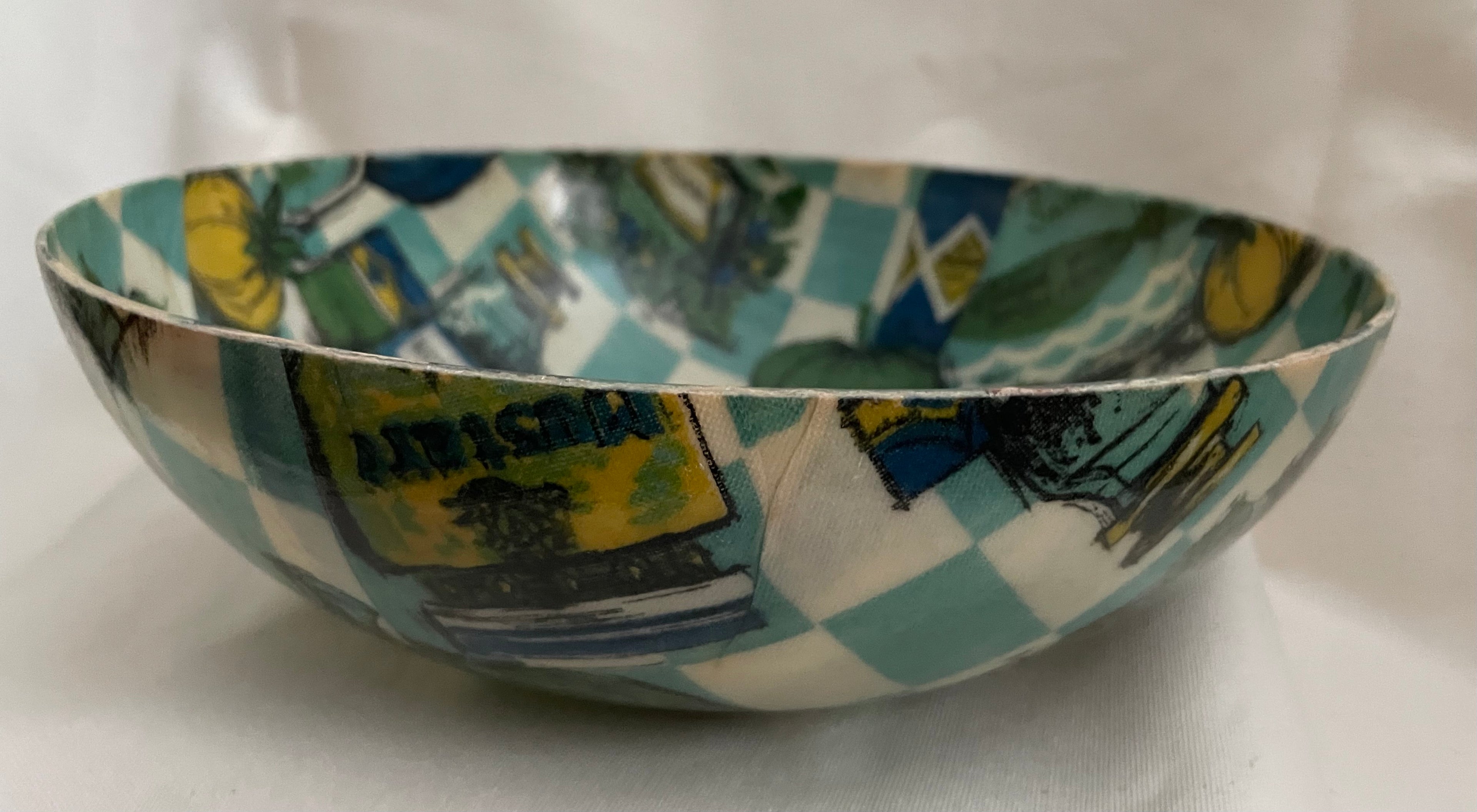 A handmade ceramic bowl featuring a colorful collage print with a teal and white checkered border and various illustrated panels, set against a plain white fabric background.