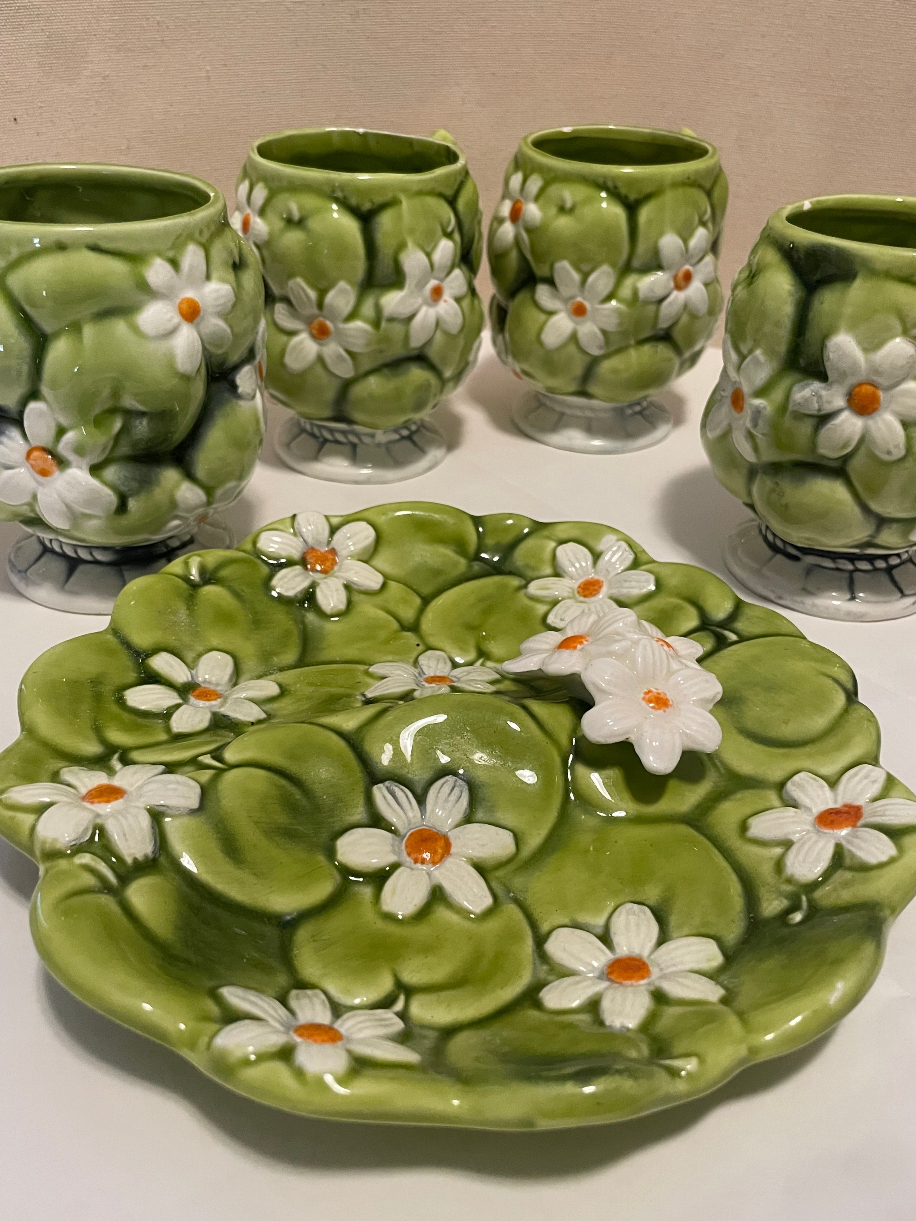A set of green ceramic tableware featuring a green apples and white daisies theme-style plate with orange centers in the foreground and four matching small coffee cups behind it.