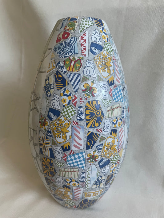 An oval ceramic vase decorated with a dense mosaic pattern of colorful French heraldic shields, fleur-de-lis, and geometric motifs in blue, gold, red, green, and white, set against a draped white fabric background.