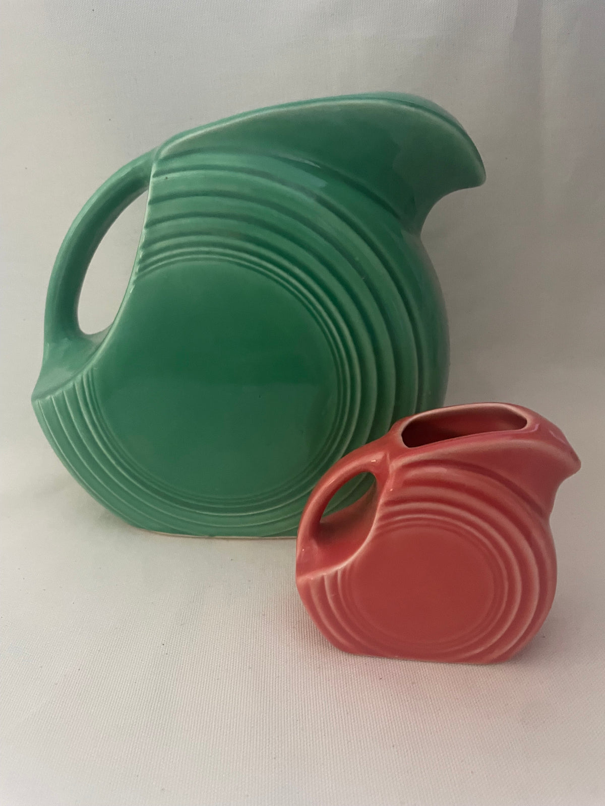 Two vintage ceramic pitchers with ribbed concentric designs, one in green and one in red, displayed on a light fabric background.