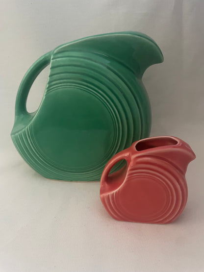 Two vintage ceramic pitchers with ribbed concentric designs, one in green and one in red, displayed on a light fabric background.