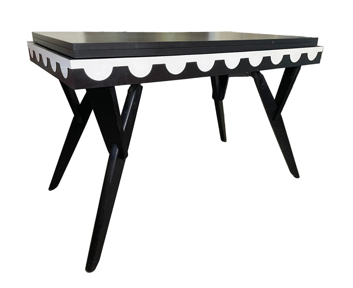 A black dining table with a white scalloped apron and angled black legs, shown in a room with gray wood-look flooring.