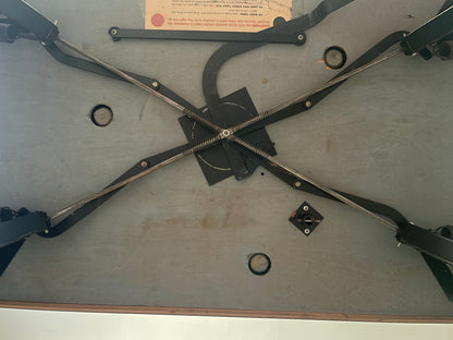 An overhead view of a vintage wooden table's underside, showing black metal support brackets and a central threaded rod assembly on a gray wood grain surface.