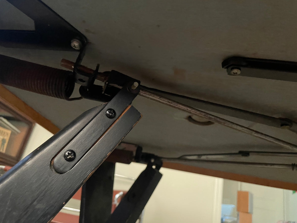 Close-up of a black metal support bracket with a spring and metal rod attached to a tabletop, showing hardware details in an indoor setting.