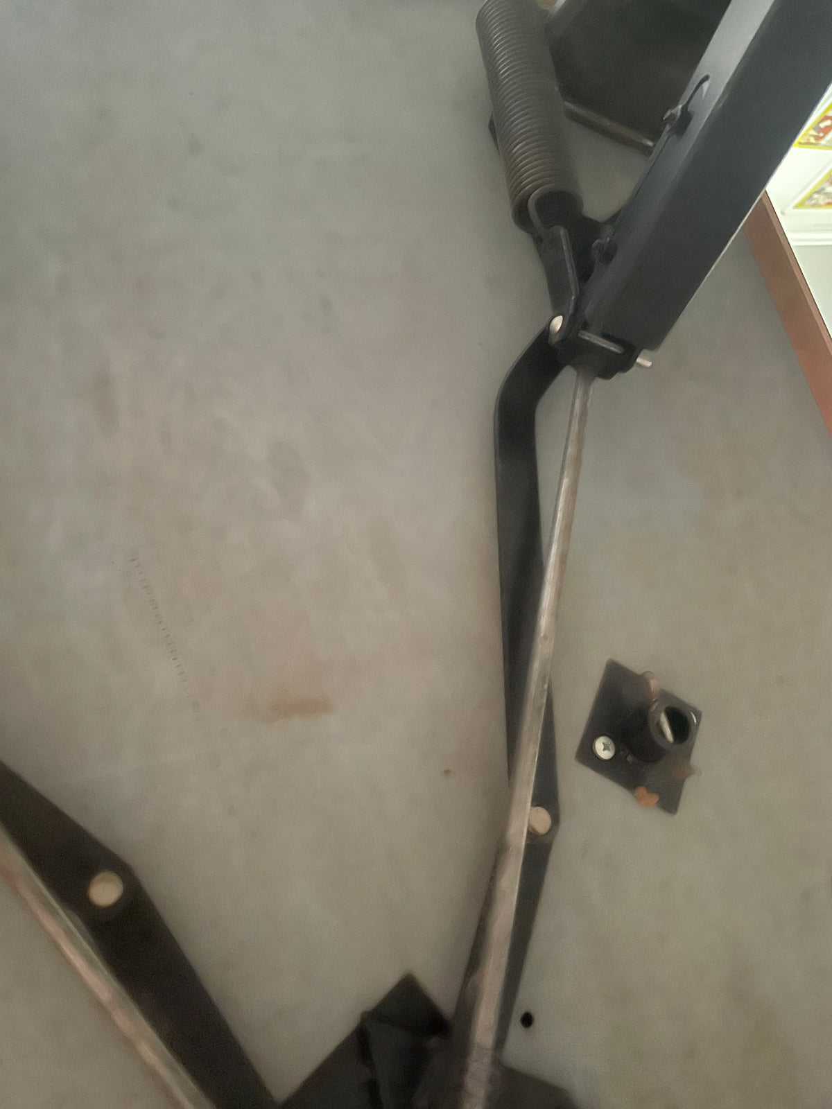 A close-up view of a black metal mechanical component with a spring and long rod on a gray concrete floor.