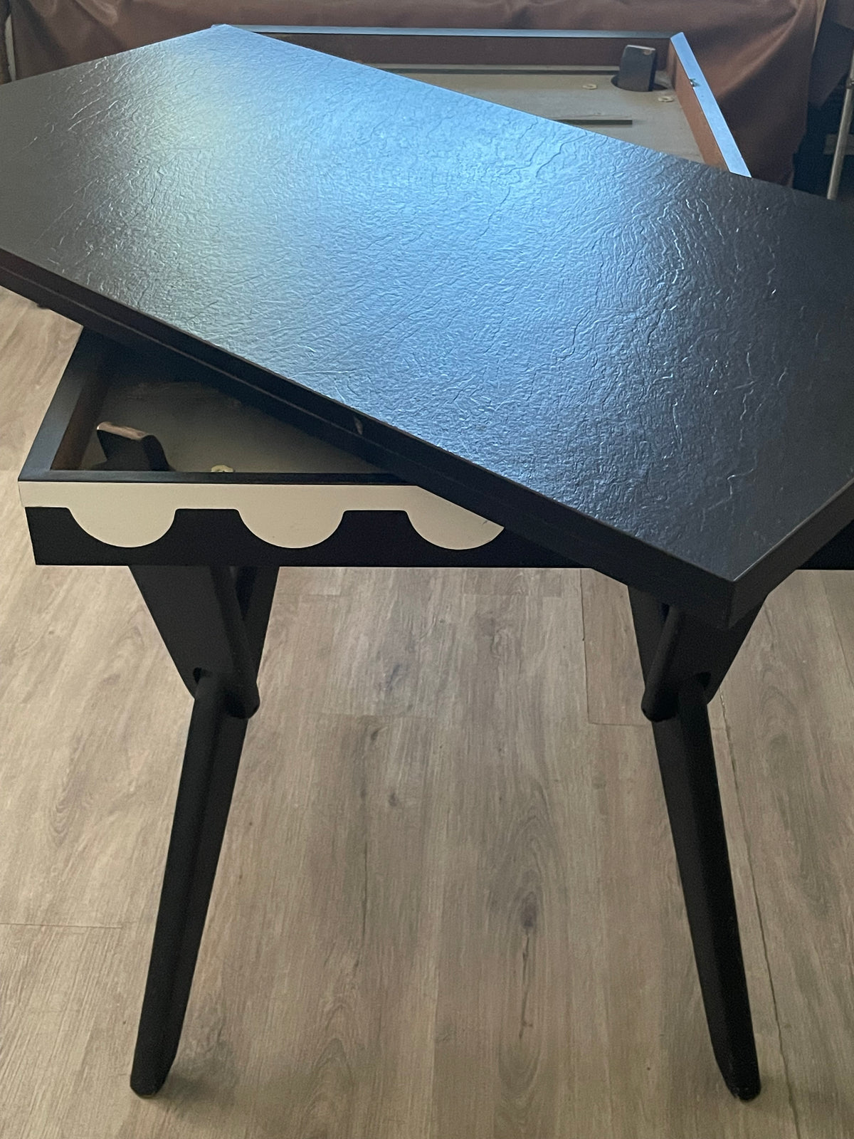 A black wooden table with a textured top and angled legs, shown with its lower drawer pulled out on a light wood floor.