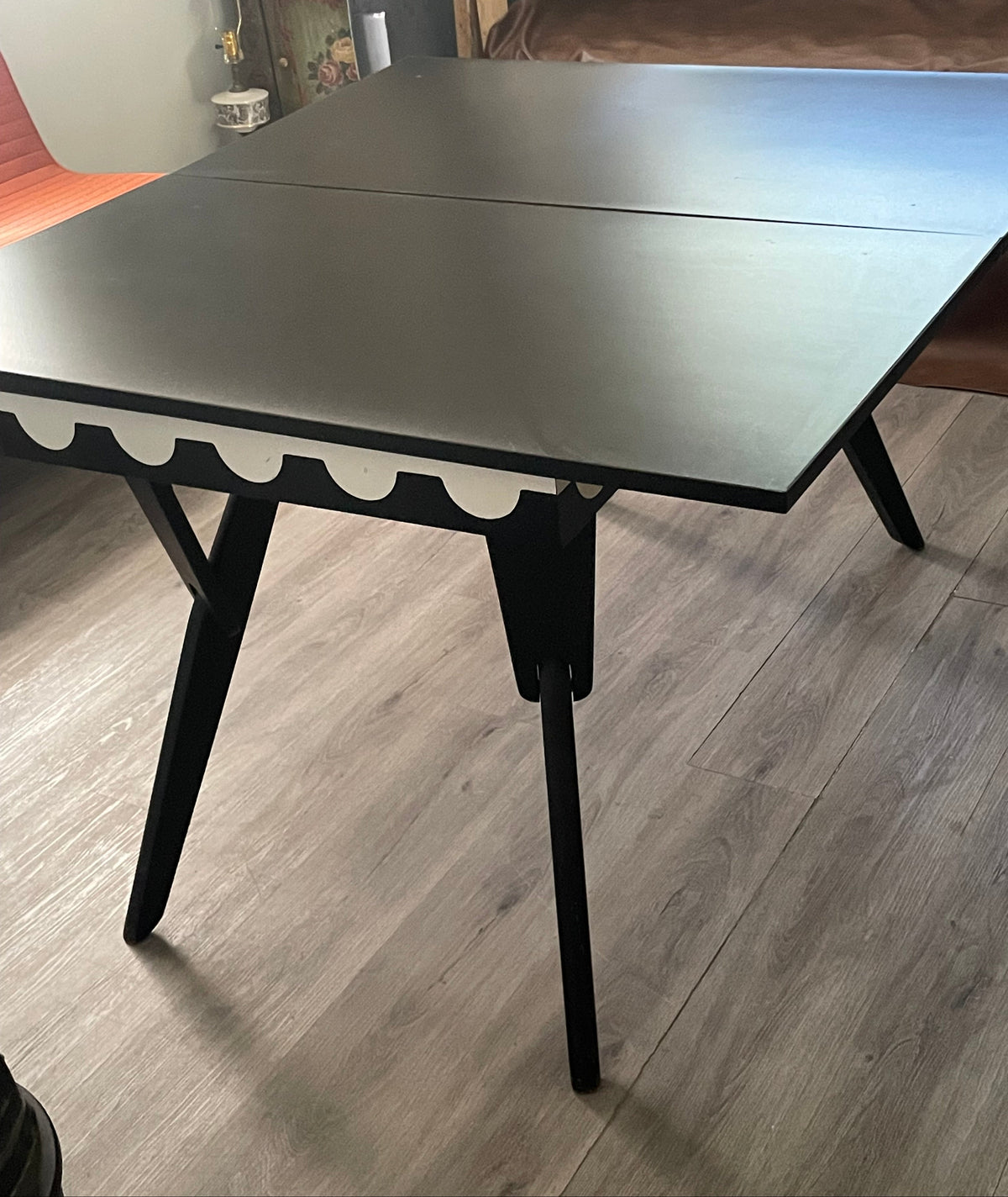 A black fold-out table with angled legs and a scalloped edge detail, shown in a home environment on a gray wood floor with a brown sofa in the background.