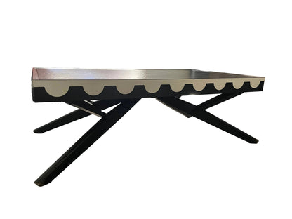 A mid-century modern coffee table with a black X-frame base and a scalloped cream top, shown in a living room setting.
