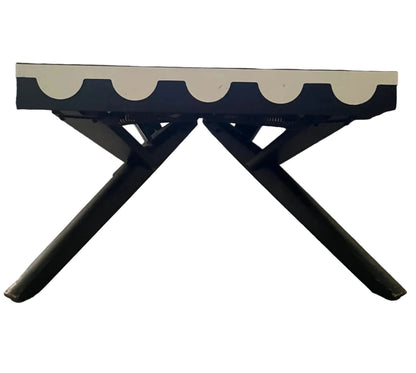 A mid-century modern lounge chair with a brown upholstered seat, a white scalloped front trim, and black metal X-style legs, set in a living room with wood floors and a bookshelf.
